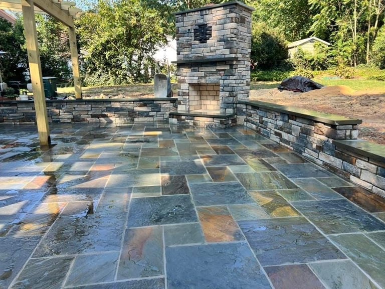 Outdoor patio with multicolored slate stone flooring, stone fireplace chimney, wooden pergola, and surrounding landscaping