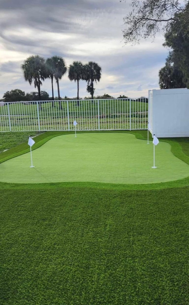 Backyard putting green with flagsticks, white fence, palm trees, and tropical landscape in background