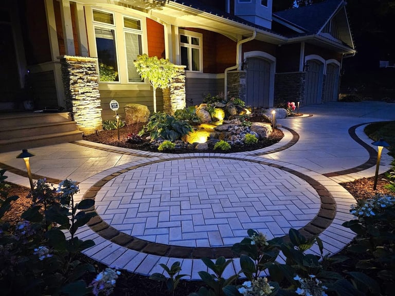 Illuminated circular paver driveway with landscape lighting at night, featuring modern home entrance and lit stone planter beds