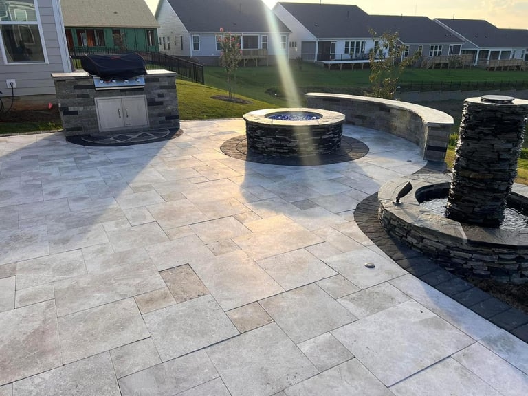 Backyard patio with stone pavers, circular fire pit, built-in seating walls, and grill under sunny afternoon light