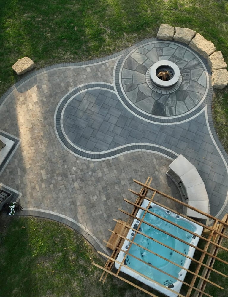 Aerial view of a curved stone patio with circular fire pit and seating area, adjacent to a swimming pool under construction