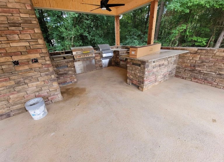Outdoor kitchen under construction with brick walls, ceiling fan, and concrete floor surrounded by green trees