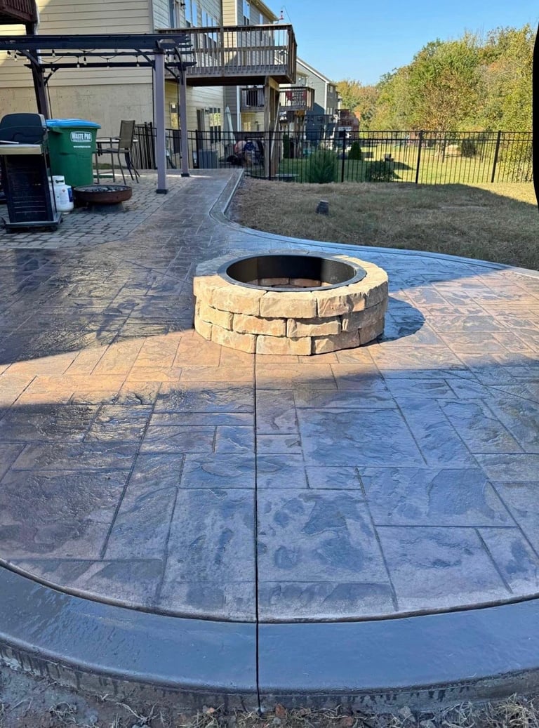 Stone fire pit on stamped concrete patio in backyard with house, deck, and fence visible
