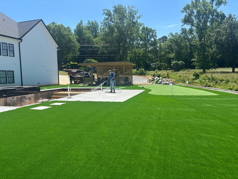 Modern backyard with bright green artificial turf, white patio area, house, and wooded background
